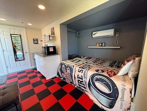 Une pièce de taille modeste avec un sol à carreaux, un lit recouvert d’une courtepointe aux motifs de flammes, une commode blanche, un four à micro-ondes et une petite fenêtre.