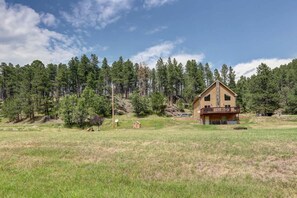A two-story log cabin with a balcony, surrounded by a forested area. - Lush Creekside Vacation Rental Haven near Mount Rushmore in South Dakota (Keystone)