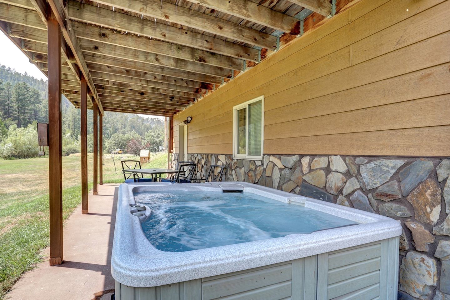 A hot tub under a wooden pergola with a stone wall and a window.