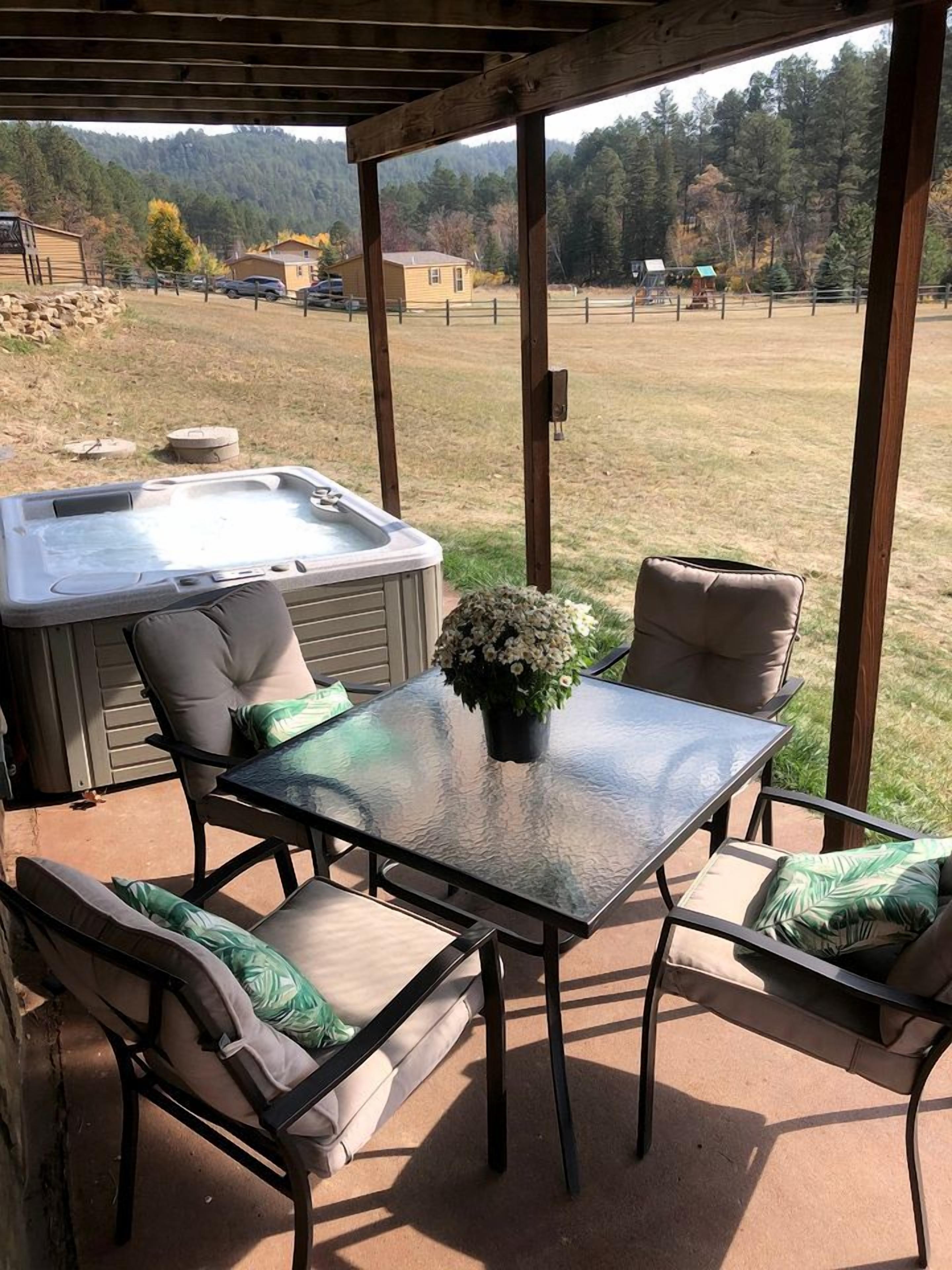 An outdoor patio with a hot tub, a glass table, and cushioned chairs.