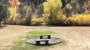Un área de picnic con dos bancos y un espacio circular de grava rodeado de césped, frente a un fondo de árboles con follaje otoñal.
