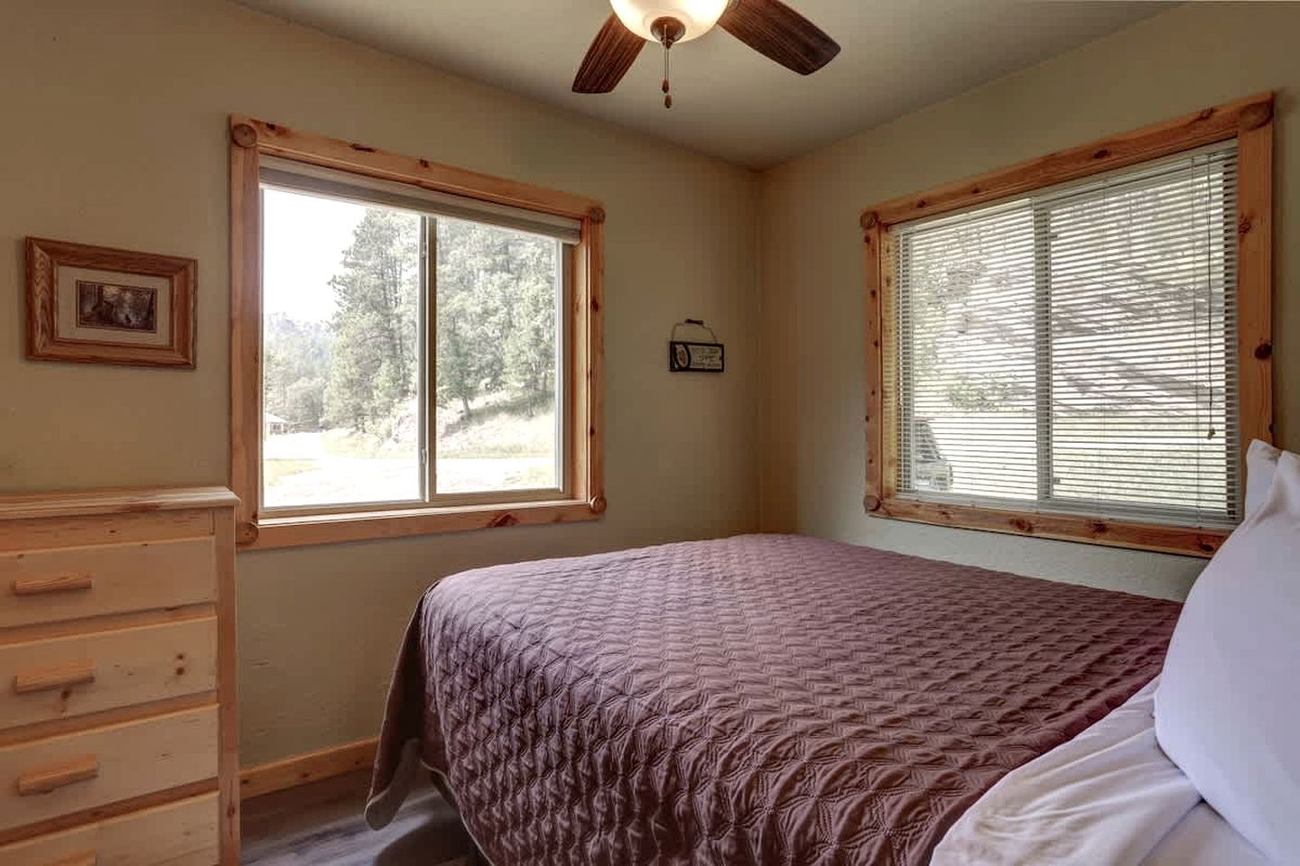 Un dormitorio con cama, cómoda, dos ventanas con persianas, ventilador de techo y un pequeño marco con un cuadro en la pared.