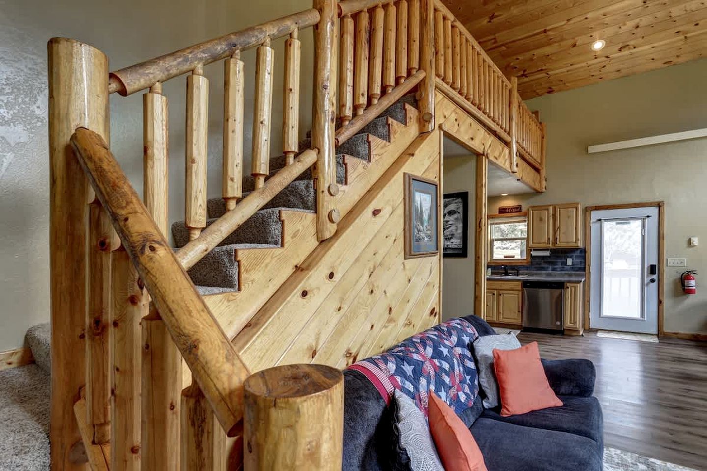 Una escalera de madera que conduce a un segundo piso, un área de cocina con armarios y una encimera, y un sofá oscuro con cojines.