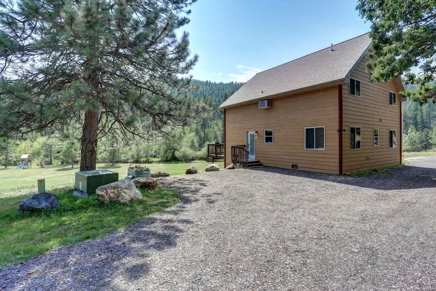 A wooden cabin with a gravel driveway, surrounded by trees and a grassy area.