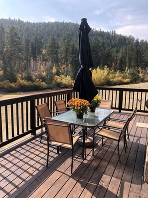 A wooden deck with a glass table and chairs, surrounded by a black railing. - Lush Creekside Vacation Rental Haven near Mount Rushmore in South Dakota (Keystone)