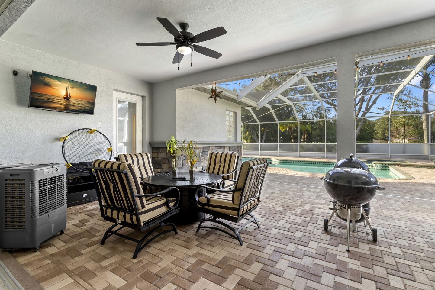 An outdoor patio with a table and chairs, a ceiling fan, a grill, and a pool in the background.