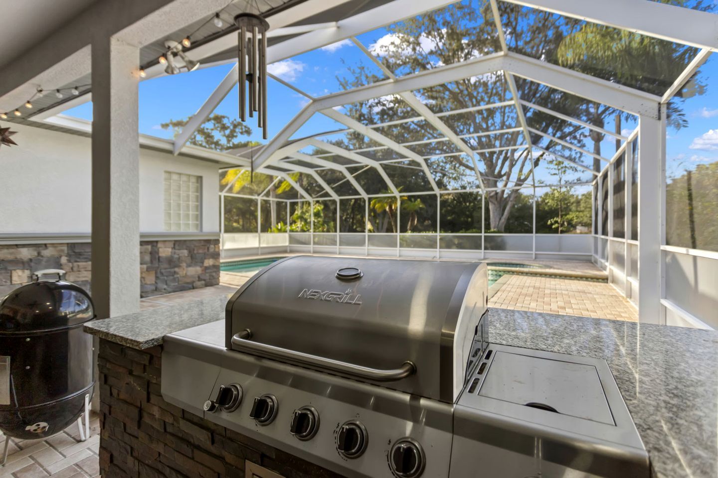 An outdoor grill area with a glass enclosure, a stone wall, and a pool in the background.