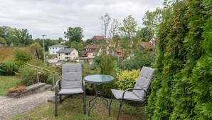 Un espacio de estar al aire libre con dos sillas y una mesa, rodeado de follaje y vistas a un barrio residencial.