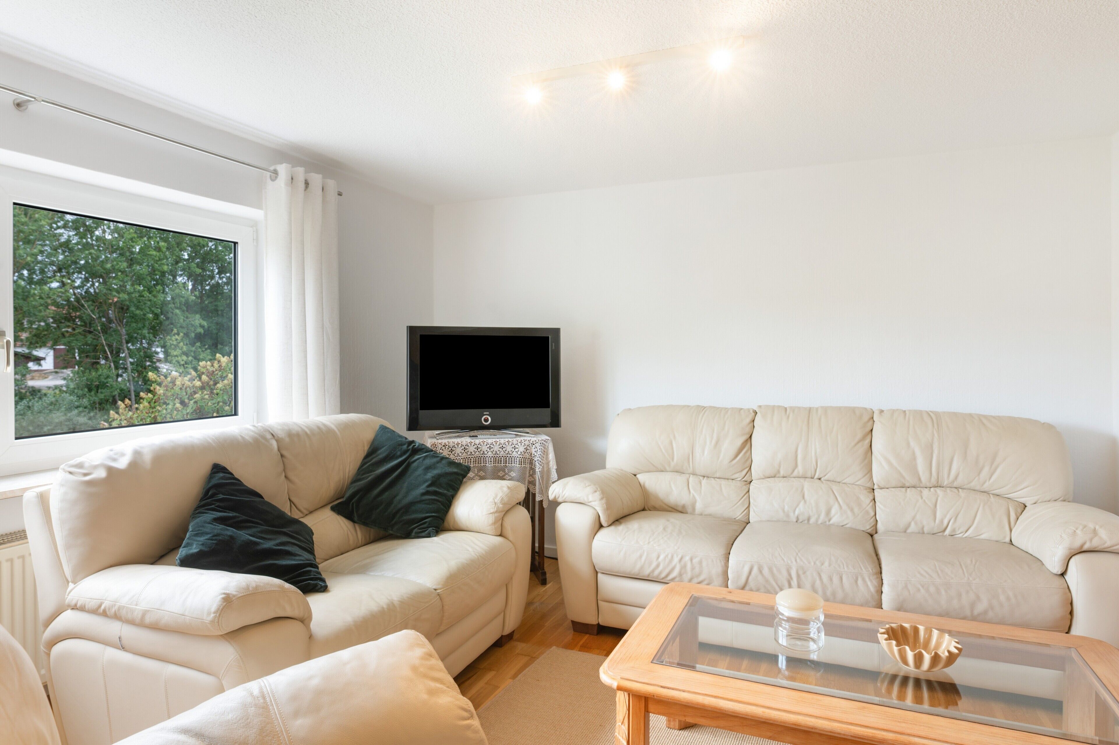 Ein Wohnzimmer mit einem beigen Sofa, einem kleinen Sessel, einem Glastisch, einem Flachbildfernseher und Blick ins Grüne.