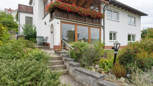 Una casa de dos pisos con un balcón, una escalera que lleva a la entrada y un jardín con diversas plantas.