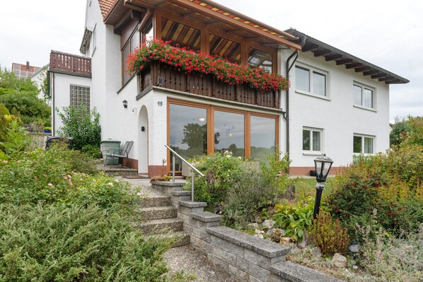 A two-story house with a balcony, a staircase leading to the entrance, and a garden with various plants.