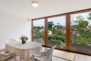 Un comedor con una mesa y sillas, un florero con flores, y vista a una zona residencial a través de amplios ventanales.