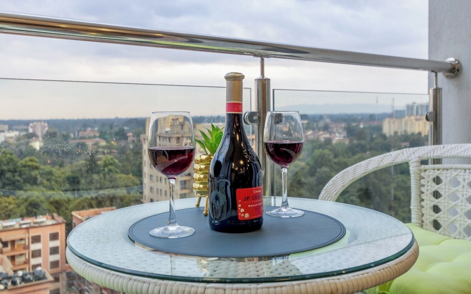 Una mesa con una botella y dos copas de vino tinto en un balcón con vista a una ciudad.