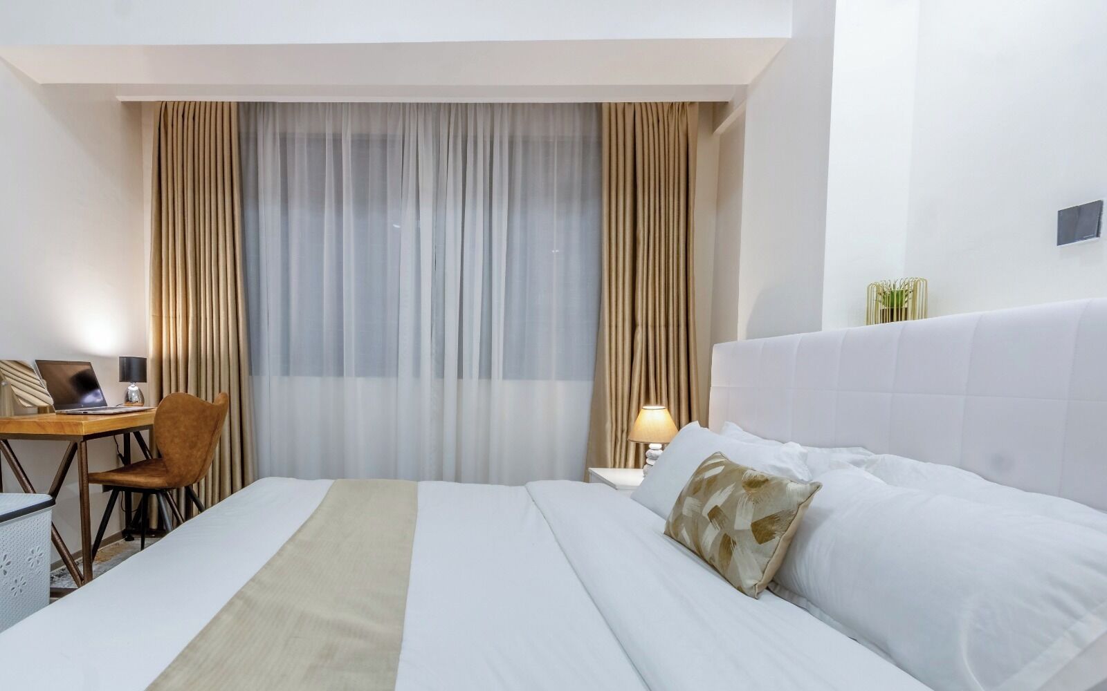 Habitación de hotel con cama, escritorio con computadora portátil, silla, mesita de noche con lámpara y cortinas.