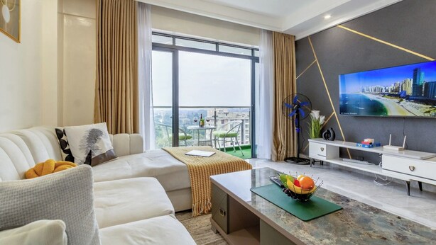 Habitación de hotel moderna con sofá, una mesa de comedor con frutero, televisor en la pared y balcón con vistas a la ciudad.