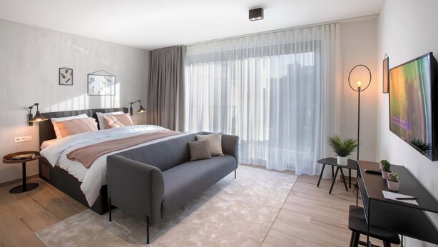 Une chambre à coucher moderne, avec un lit, des tables de chevet, un bureau, une chaise et une grande fenêtre ornée de rideaux légers.