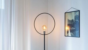 Une lampe de sol avec un abat-jour circulaire et une ampoule allumée, un tableau encadré accroché au mur et une fenêtre avec des rideaux blancs.