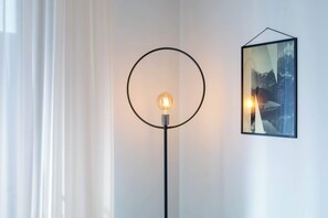 Una làmpada de peu amb pantalla circular i una bombeta encèsa, un quadre amb marc a la paret, i una finestra amb cortines blanques.