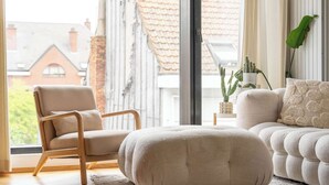A modern living room with a wooden chair, a cushioned ottoman, a sofa, and a view of buildings through large windows. - Bright Duplex with Balcony in Antwerp (Antwerpen)