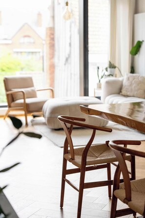 Uma sala de estar moderna com uma mesa de jantar de madeira, duas cadeiras de vime, um sofá e vista para uma casa por uma janela grande.