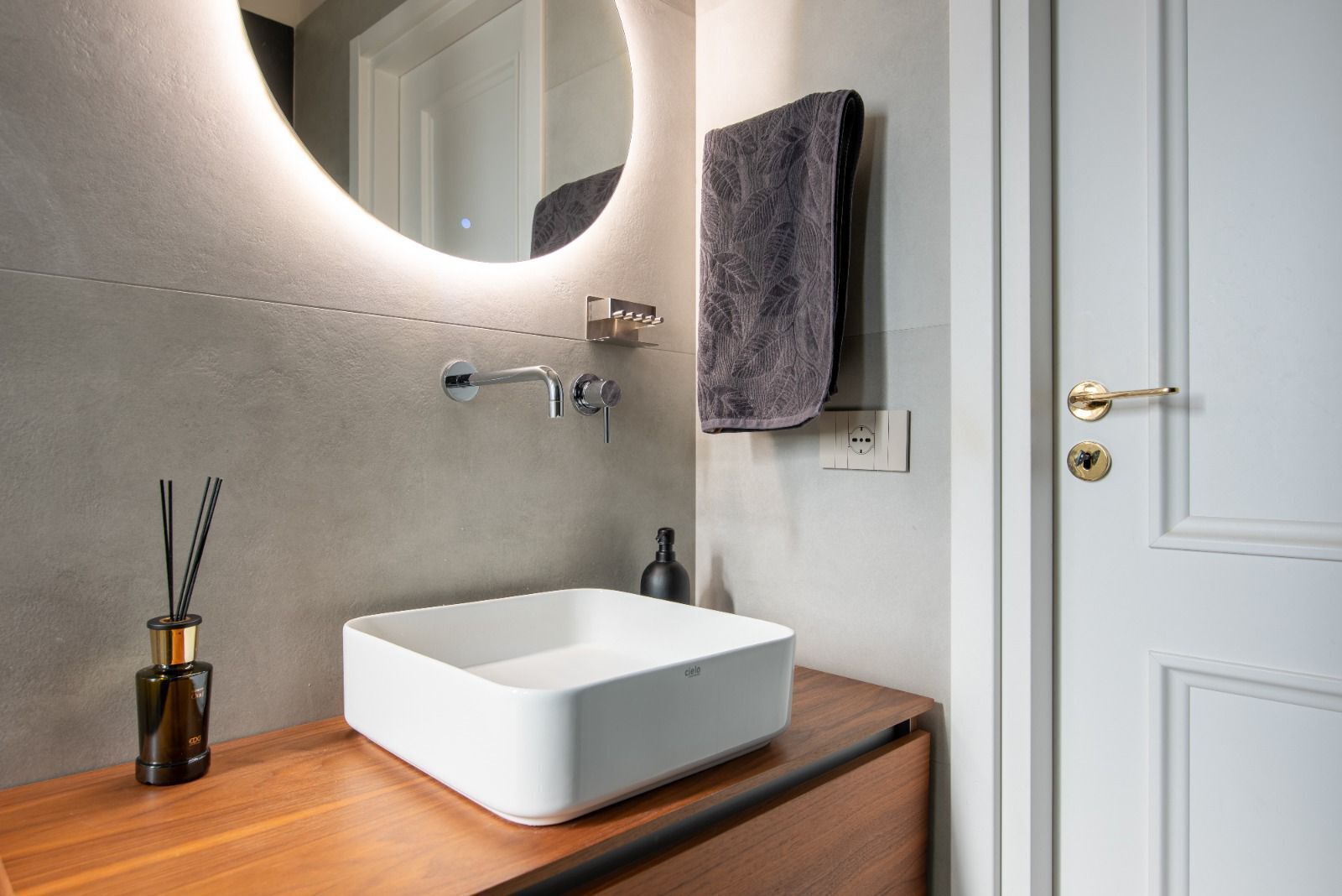 Une salle de bain avec un lavabo blanc, un miroir doté d’un éclairage circulaire, une serviette et une porte.
