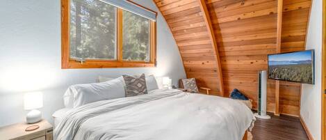 A bedroom with a large bed, a window, a TV, and a wooden ceiling.