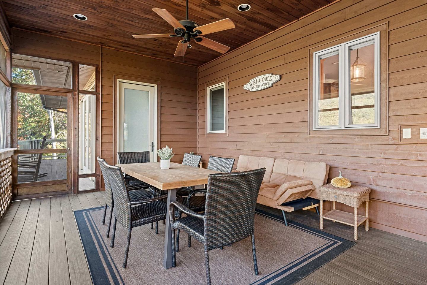 A wooden porch with a table and chairs, a ceiling fan, and a sign that reads 'WELCOME TO OUR PORCH.'