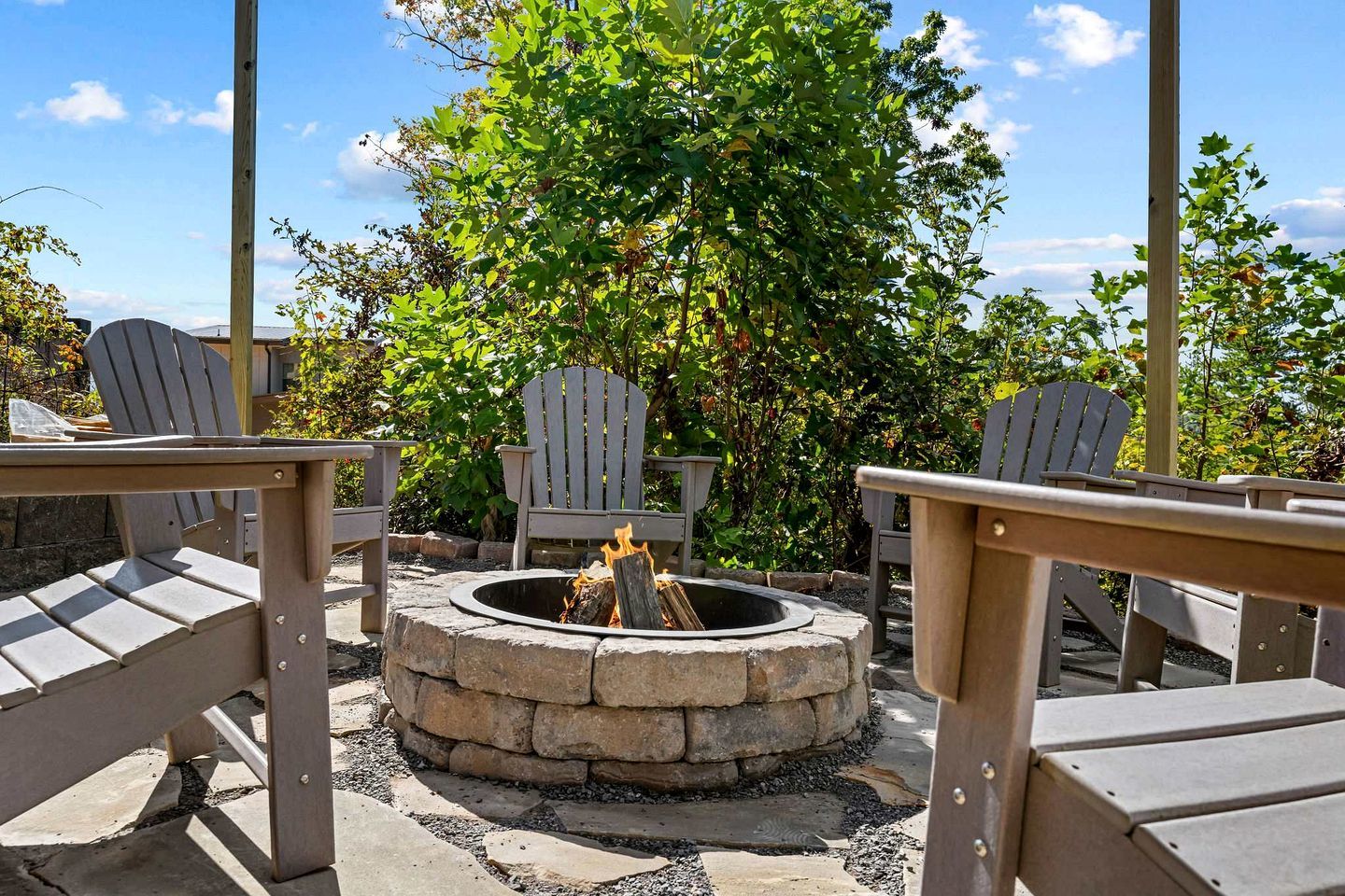 An outdoor seating area with Adirondack chairs, a stone fire pit, and a backdrop of trees and shrubs.