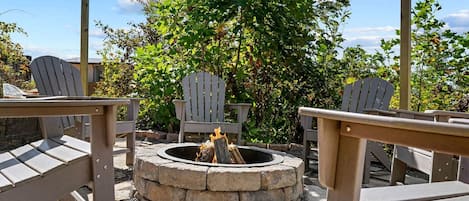 An outdoor seating area with Adirondack chairs, a stone fire pit, and a backdrop of trees and shrubs.