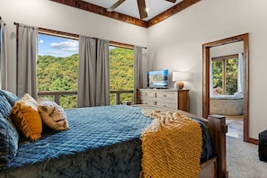 A bedroom with a large bed, a television, a dresser, and a view of a forest through large windows. - Luxury Mountain Cabin with Hot Tub and Panoramic Views near Pigeon Forge, Tennessee (Sevierville)