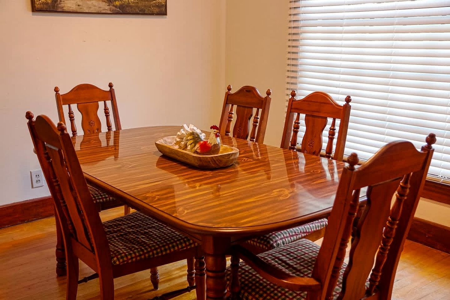 Comedor con mesa y sillas de madera, un cuenco con comida y una decoración de gallo, además de una ventana con persianas.