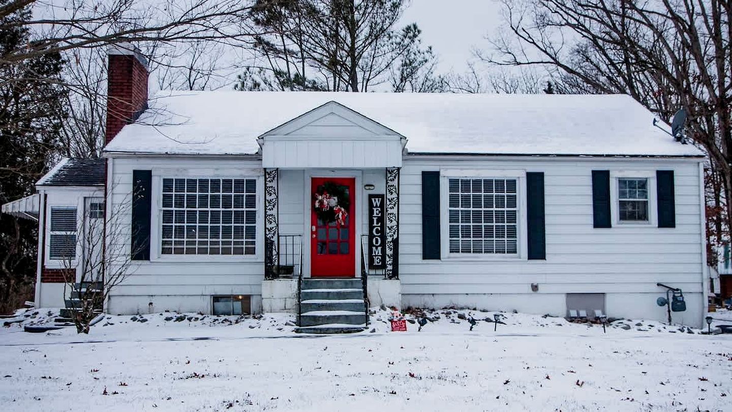 Ein weißes Haus mit roter Tür, schwarzen Fensterläden und einem mit Schnee bedeckten Dach.