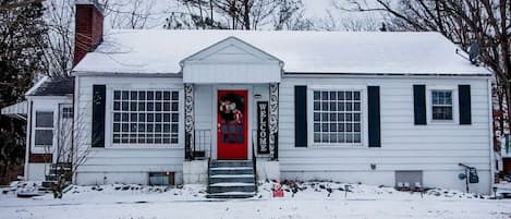 A white house with a red door, black shutters, and a snow-covered roof.