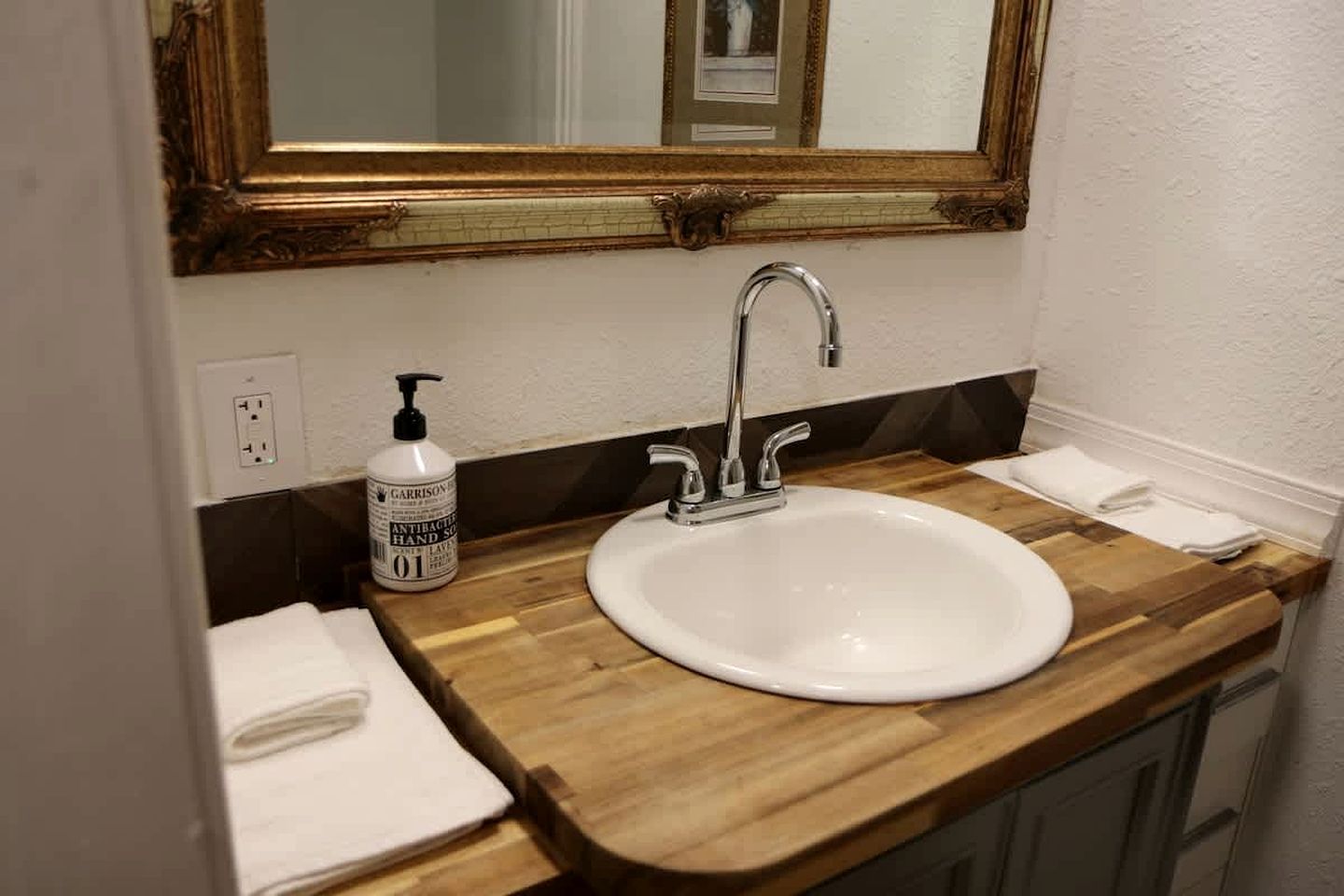 A bathroom with a wooden vanity, a white sink, a mirror with a gold frame, a bottle of hand soap, and a towel.