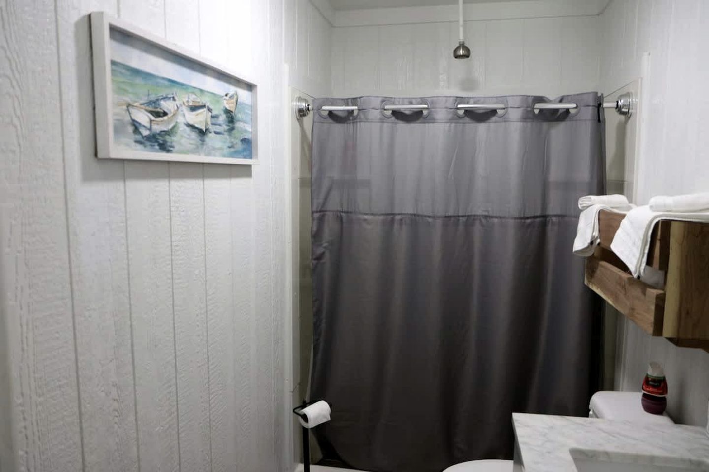 A bathroom with a grey shower curtain, a painting of boats, a towel rack, and a small wooden shelf with a bottle.