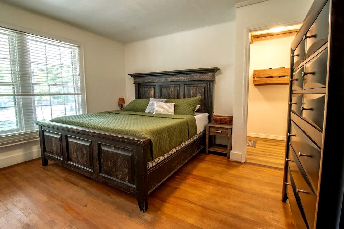 A bedroom with a wooden bed, a nightstand, a dresser, and a window with blinds.