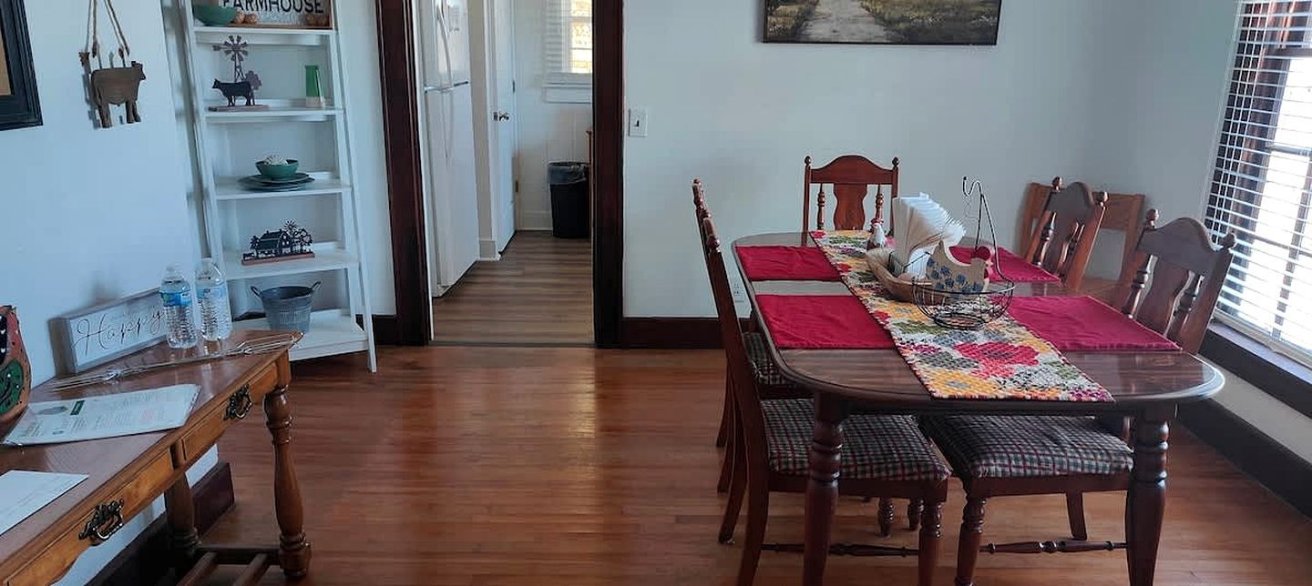 Un coin repas avec une table en bois, des chaises et un chemin de table rouge. On y trouve une bibliothèque avec des objets décoratifs et une fenêtre avec des stores.