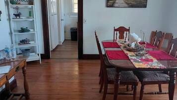 Un coin repas avec une table en bois, des chaises et un chemin de table rouge. On y trouve une bibliothèque avec des objets décoratifs et une fenêtre avec des stores.