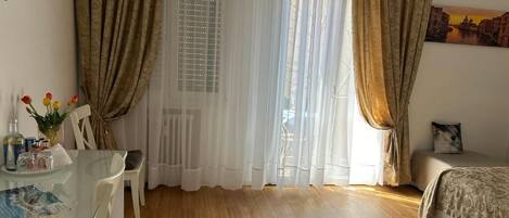 A room with a bed, a glass table with a vase, curtains, and a painting on the wall.