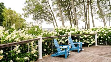 En træterrasse med to blå Adirondack-stole, omgivet af hvide blomster og træer.