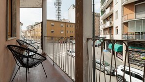 A balcony with metal railings, a view of a street with parked cars, and a building with balconies and a shop. - A modern and welcoming 55-square-meter apartment, ideal for up to 4 guests, located on the first floor of a building with an elevator. The property is situated in Brenta, a quiet residential area a short distance from Porta Romana and the new Scalo Roman (Milan)