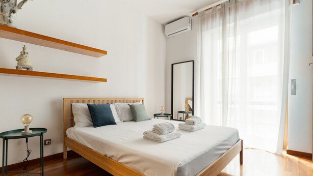 A neatly arranged bedroom with a wooden bed, bedside tables, a mirror, and a window with sheer curtains. - A modern and welcoming 55-square-meter apartment, ideal for up to 4 guests, located on the first floor of a building with an elevator. The property is situated in Brenta, a quiet residential area a short distance from Porta Romana and the new Scalo Roman (Milan)