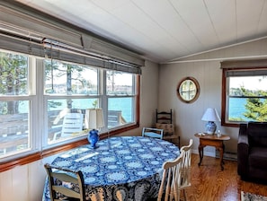 Ein Zimmer mit einem runden Tisch, der mit einer gemusterten Tischdecke bedeckt ist, einer blauen Vase und einem Holzstuhl. Es gibt ein Fenster mit Blick ins Freie, einen runden Spiegel an der Wand und einen kleinen Tisch mit einer Lampe.
