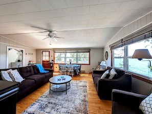 Ein geräumiges Wohnzimmer mit Deckenventilator, eine Essecke mit Tisch und Stühlen sowie große Fenster mit Blick ins Freie.