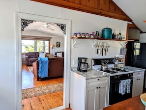 Eine Küche mit Herd, Kaffeemaschine und verschiedenen Küchenutensilien. Ein Esstisch und ein Sofa befinden sich im angrenzenden Wohnzimmer.