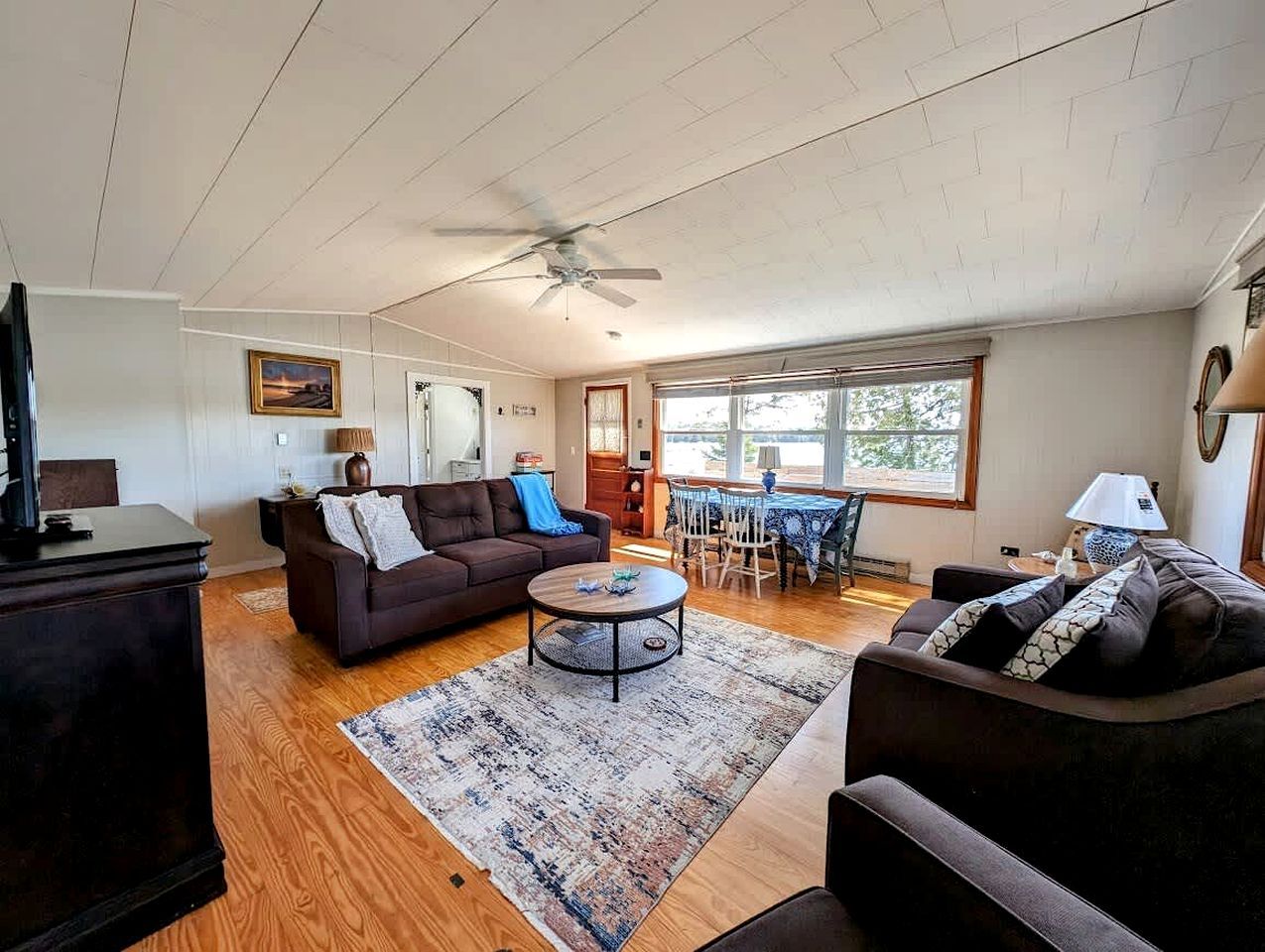 A spacious living room with a ceiling fan, a dining area with a table and chairs, and a large window providing natural light.