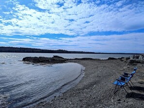 Ein Strand mit einer Reihe blauer Stühle, die zum Wasser zeigen, einer felsigen Küste und einem bewaldeten Horizont.