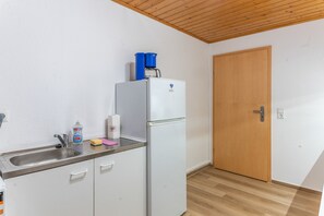 A small kitchenette with a white refrigerator, a coffee maker, a sink with a faucet, and a wooden door.