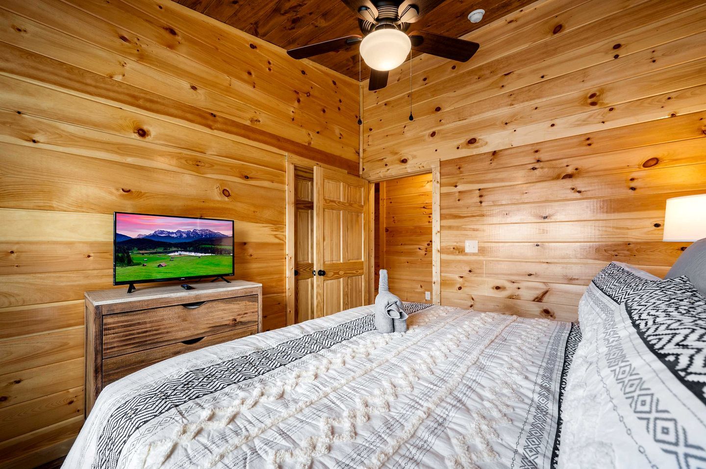 A wooden cabin interior with a bed, a television on a dresser, and a ceiling fan.