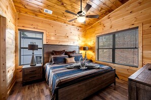 Une chambre à coucher dans une cabane en bois, comprenant un lit, deux tables de chevet, un ventilateur de plafond et deux fenêtres équipées de stores.